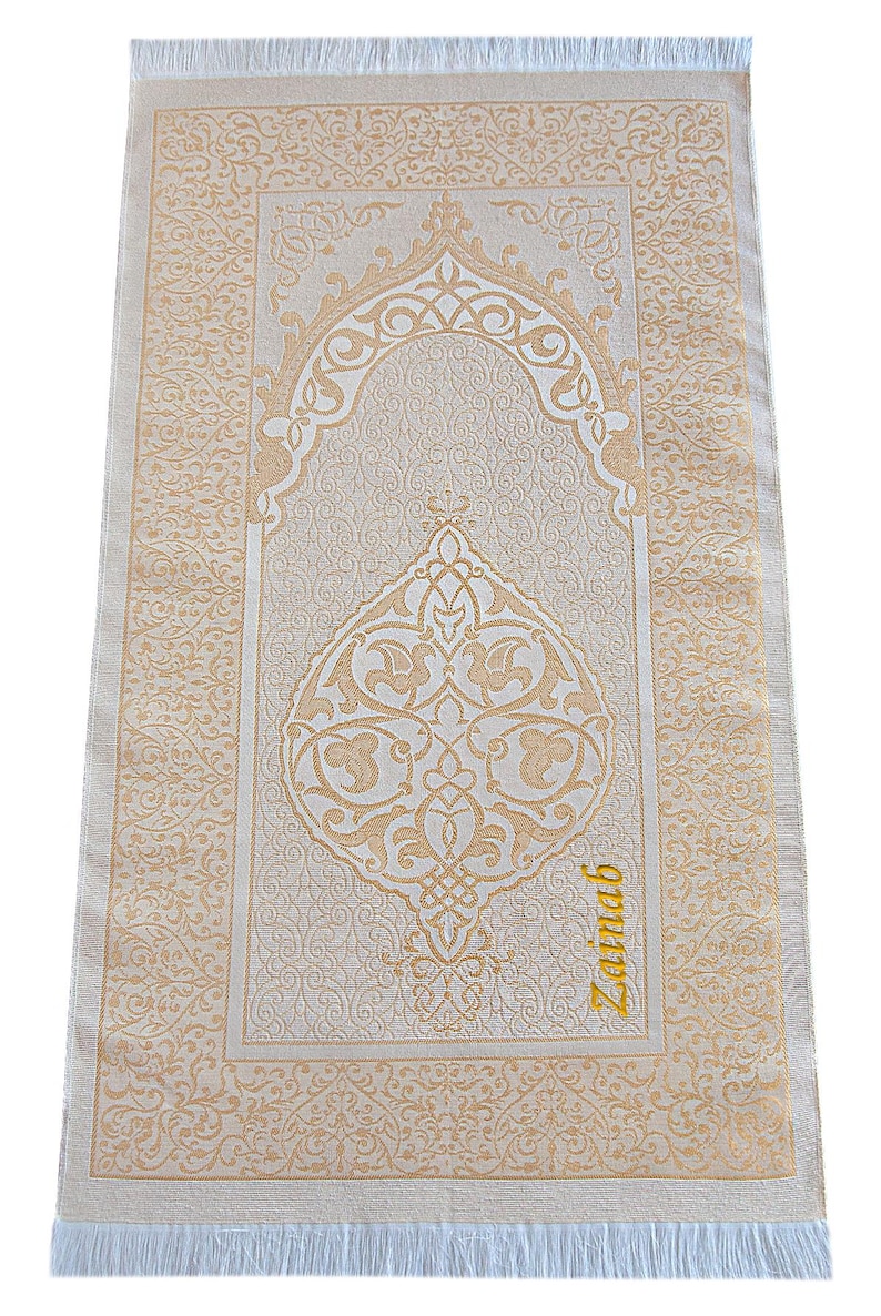 May include: Beige prayer rug with intricate gold-colored floral and geometric patterns. The rug features a central arched design and a decorative border. The word "Zainab" is written in gold script on the bottom right corner. The rug has a fringed edge.