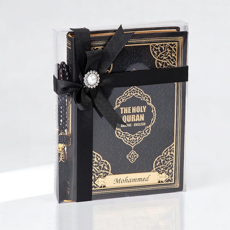 May include: A boxed set featuring a black Quran with gold filigree, a prayer bead, and a black ribbon with a pearl-centered embellishment. The Quran is labeled "THE HOLY QURAN Arabic - English" with the name "Mohammed" below.
