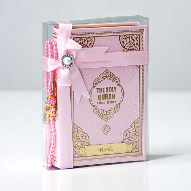 May include: A pink Quran with gold accents and the text "THE HOLY QURAN ARABIC-ENGLISH" is presented in a clear box. A pink ribbon with a pearl embellishment and a matching pink prayer beads complete the set. The name "Maida" is printed on the cover.