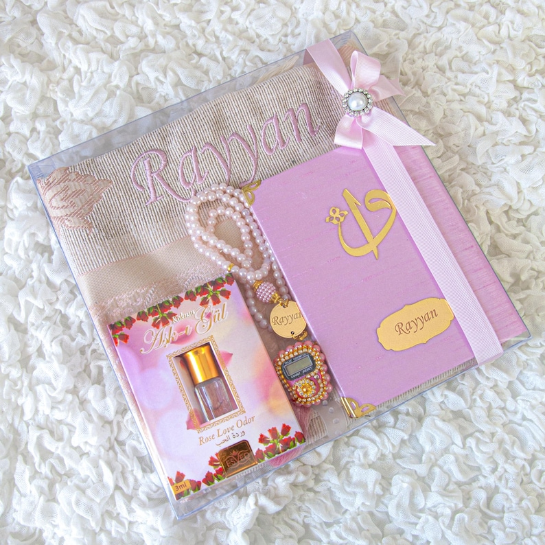 May include: A gift set in a clear box, featuring a prayer mat embroidered with "Rayyan", a pink book with gold accents, a pearl necklace, a perfume bottle, and a beaded counter. The set is tied with a pink ribbon and bow.