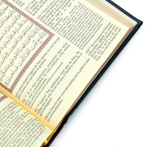 May include: Open book with Arabic script and French translation. The pages are cream-colored with black text and red accents. The book has a black cover and is open on a white surface.