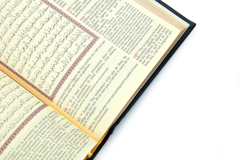 May include: Open book with Arabic script and French translation. The pages are cream-colored with black text and red accents. The book has a black cover and is open on a white surface.