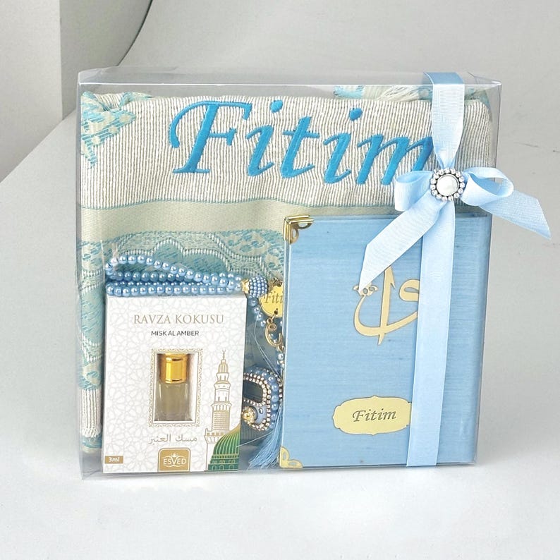 May include: A gift set in a clear box, featuring a light blue prayer rug with "Fitim" embroidered in blue, a blue book with gold accents, a bottle of perfume, and a string of beads. A light blue ribbon with a decorative clasp secures the box.