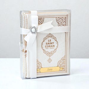 May include: A white book with gold accents, titled "LE SAINT CORAN ARABE FRAN&Ccedil;AIS", is packaged in a clear box and tied with a white ribbon and a decorative clasp. A string of pearls is visible on the left. The name "Sara" is printed on a gold plate.