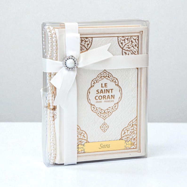 May include: A white book with gold accents, titled "LE SAINT CORAN ARABE FRAN&Ccedil;AIS", is packaged in a clear box and tied with a white ribbon and a decorative clasp. A string of pearls is visible on the left. The name "Sara" is printed on a gold plate.