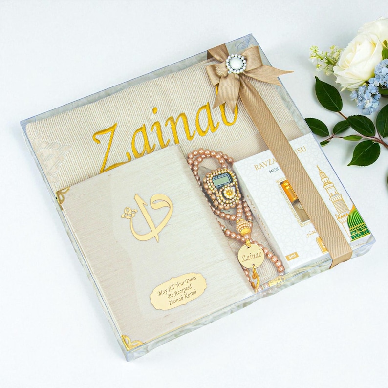 May include: A gift set in a clear box, featuring a beige prayer mat with gold embroidery, a book, a beaded rosary, and a small perfume box. The set is tied with a beige ribbon and a decorative bow.