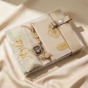 May include: A gift set in a clear box, featuring a beige prayer rug embroidered with "Ayla" in gold, a matching book, and a beaded rosary. The book is adorned with a gold embellishment and a ribbon with a pearl accent.