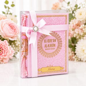 Op de afbeelding: Een roze Al-Qur'an Al-Karim boek met gouden letters en een decoratieve rand, verpakt in een doorzichtige plastic doos. Het boek is omwikkeld met een lichtroze lint met een parelmoer ornament en bevat roze gebedskralen.