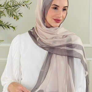 May include: A sheer hijab in shades of gray and beige with a striped pattern. The headscarf is draped over the head and shoulders. The person is wearing a white blouse.