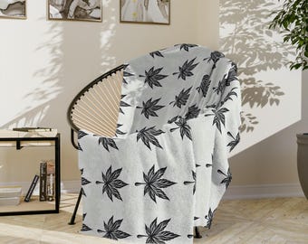 May include: A white throw blanket draped over a chair, featuring a repeating pattern of black maple leaves. The blanket is made of a soft material and adds a decorative touch to the room. The chair has a black metal frame and a woven back.