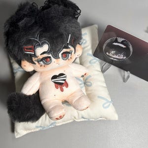 May include: A plush toy with black hair, red eyes, and a black and white heart on its chest. The toy is lying on a white pillow with a blue ribbon pattern. The toy has a black tail and is wearing a black lace headband.