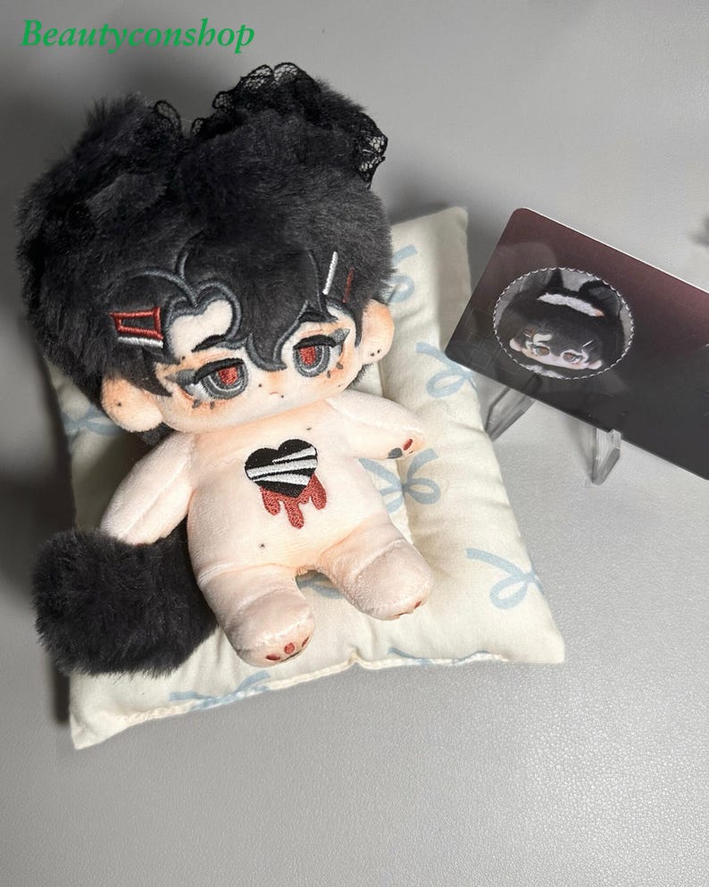 May include: A plush toy with black hair, red eyes, and a black and white heart on its chest. The toy is lying on a white pillow with a blue ribbon pattern. The toy has a black tail and is wearing a black lace headband.