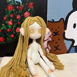 May include: A handmade doll with long, blonde yarn hair, button eyes, and a white fabric body. The doll is sitting on a white fabric surface.