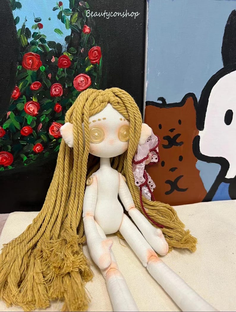 May include: A handmade doll with long, blonde yarn hair, button eyes, and a white fabric body. The doll is sitting on a white fabric surface.