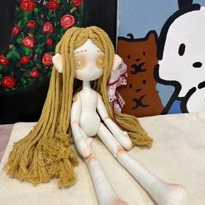 May include: A handmade doll with a white body, long blonde hair made of rope, and large, round, yellow eyes. The doll is sitting on a white fabric surface.