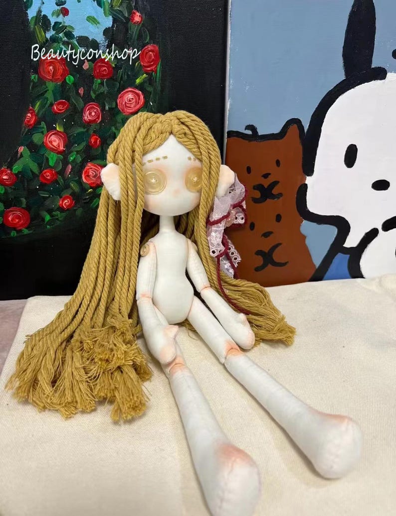 May include: A handmade doll with a white body, long blonde hair made of rope, and large, round, yellow eyes. The doll is sitting on a white fabric surface.