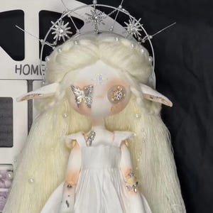 May include: A white doll with long blonde hair wearing a white dress and a silver crown with star-shaped embellishments. The doll has large, round eyes with butterfly and star-shaped decorations.