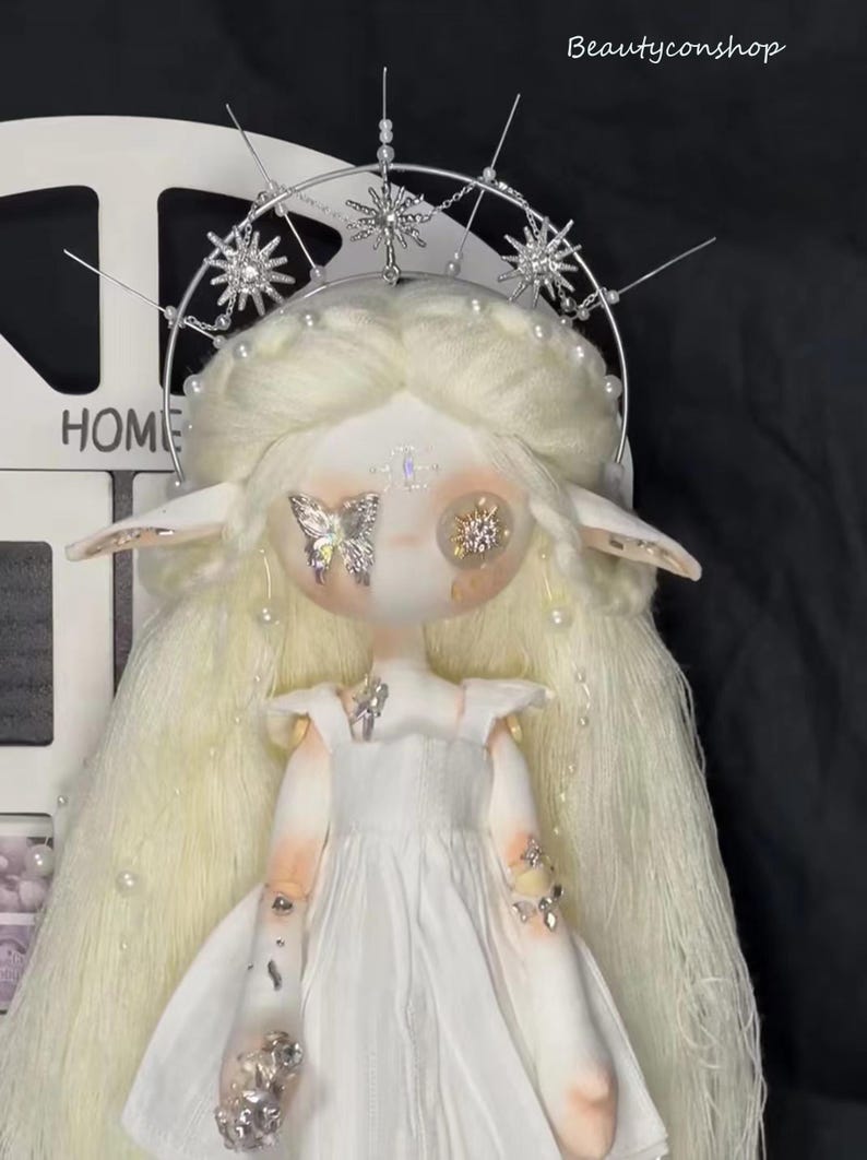 May include: A white doll with long blonde hair wearing a white dress and a silver crown with star-shaped embellishments. The doll has large, round eyes with butterfly and star-shaped decorations.