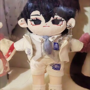 May include: A plush toy doll with black hair, wearing a white jacket, a striped tie, and white pants. The doll has a white and blue design on the pants and a silver chain necklace with a pendant.