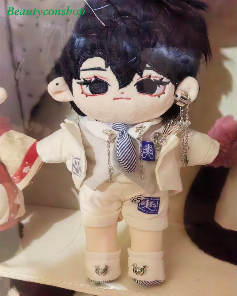 May include: A plush toy doll with black hair, wearing a white jacket, a striped tie, and white pants. The doll has a white and blue design on the pants and a silver chain necklace with a pendant.