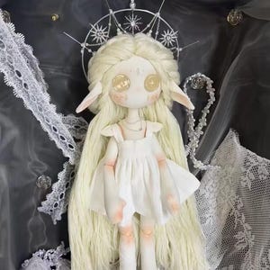 May include: A white porcelain doll with long blonde hair wearing a white dress and a silver crown with star-shaped accents. The doll has large, golden eyes and pointed ears.