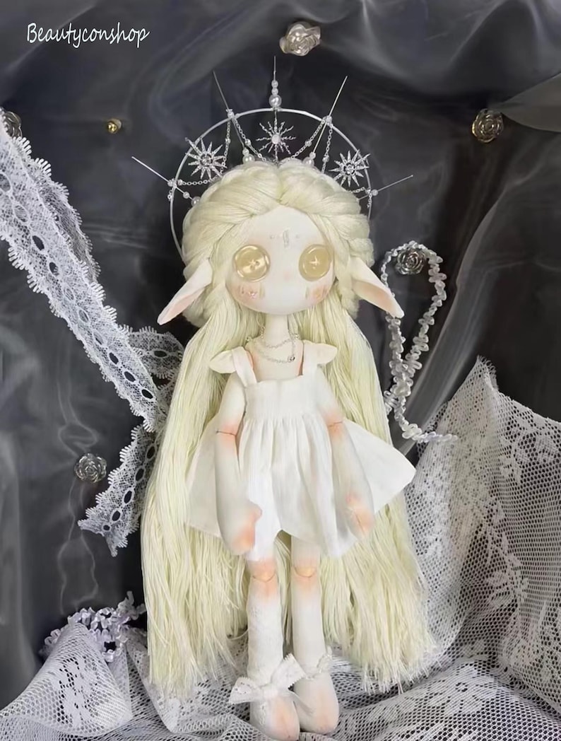 May include: A white porcelain doll with long blonde hair wearing a white dress and a silver crown with star-shaped accents. The doll has large, golden eyes and pointed ears.