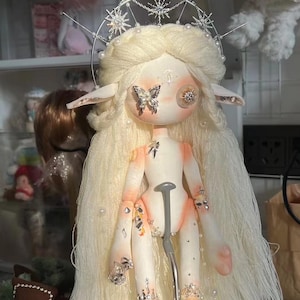 May include: A white doll with long blonde hair and a silver crown. The doll has large, round eyes and a butterfly on one eye. The doll is wearing a silver crown with stars and pearls. The doll is decorated with rhinestones and pearls.