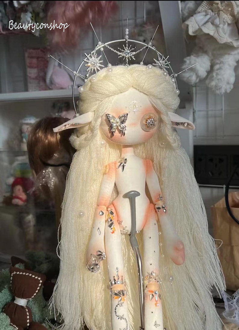 May include: A white doll with long blonde hair and a silver crown. The doll has large, round eyes and a butterfly on one eye. The doll is wearing a silver crown with stars and pearls. The doll is decorated with rhinestones and pearls.