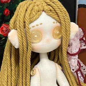 May include: A handmade doll with long, braided, yellow yarn hair. The doll has a white fabric body with button eyes and a button on each shoulder. The doll is wearing a white fabric dress with pink and white lace trim.