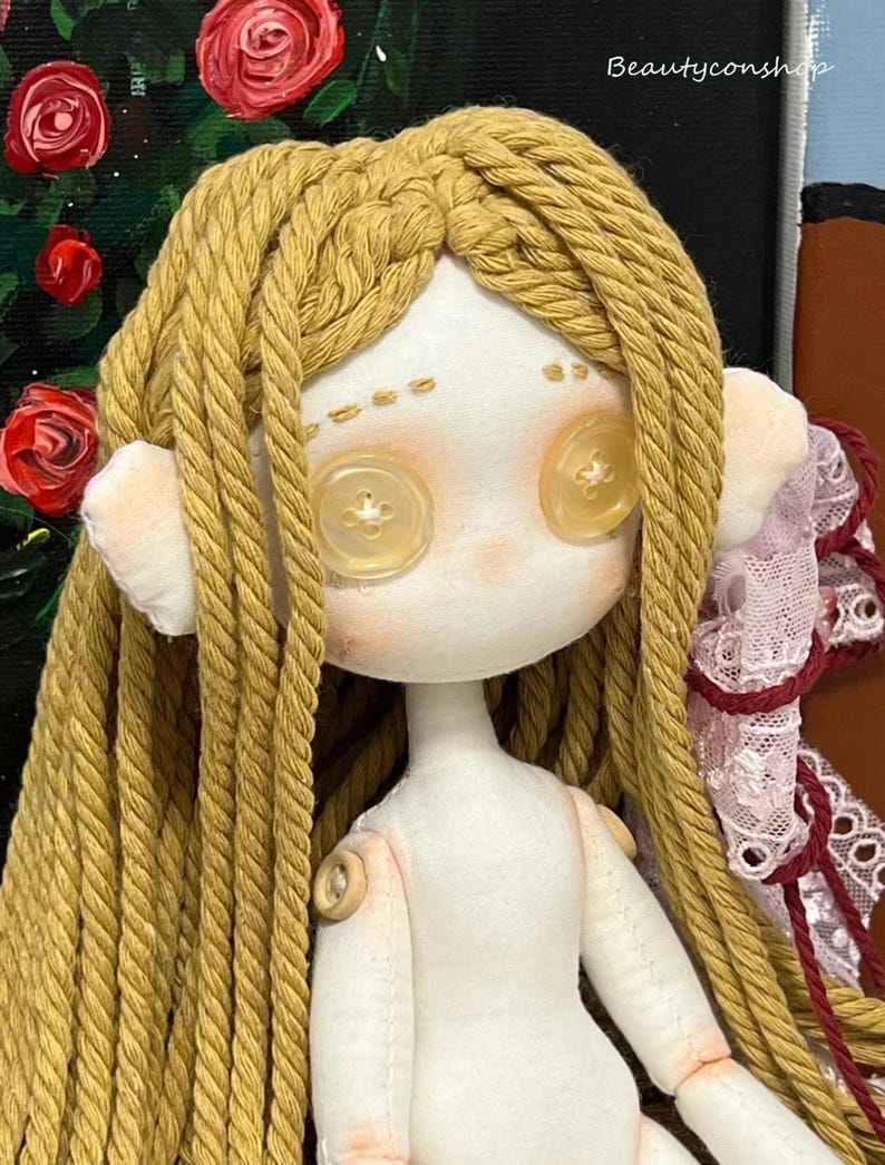 May include: A handmade doll with long, braided, yellow yarn hair. The doll has a white fabric body with button eyes and a button on each shoulder. The doll is wearing a white fabric dress with pink and white lace trim.