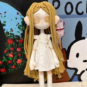 May include: A handmade doll with long blonde yarn hair, a white dress, and button eyes. The doll is standing on a white surface.