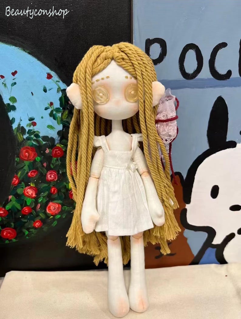 May include: A handmade doll with long blonde yarn hair, a white dress, and button eyes. The doll is standing on a white surface.