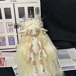 May include: A white porcelain doll with long white hair and a silver crown. The doll is wearing a white dress with rhinestones and has a butterfly on its face. The doll is sitting on a white surface.