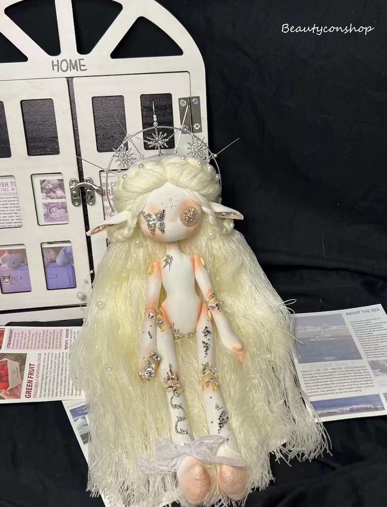 May include: A white porcelain doll with long white hair and a silver crown. The doll is wearing a white dress with rhinestones and has a butterfly on its face. The doll is sitting on a white surface.