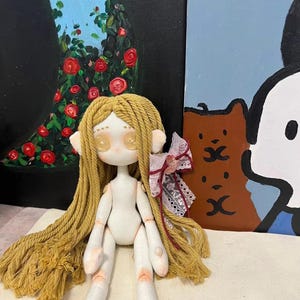 May include: A handmade doll with long blonde yarn hair, white fabric body, and painted details. The doll has large, round eyes with painted eyelashes and a pink fabric bow on its head. The doll is sitting on a white fabric surface.