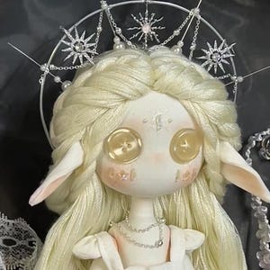 May include: A white porcelain doll with long blonde hair wearing a white dress and a silver crown with pearl accents. The doll has large, round, yellow button eyes.