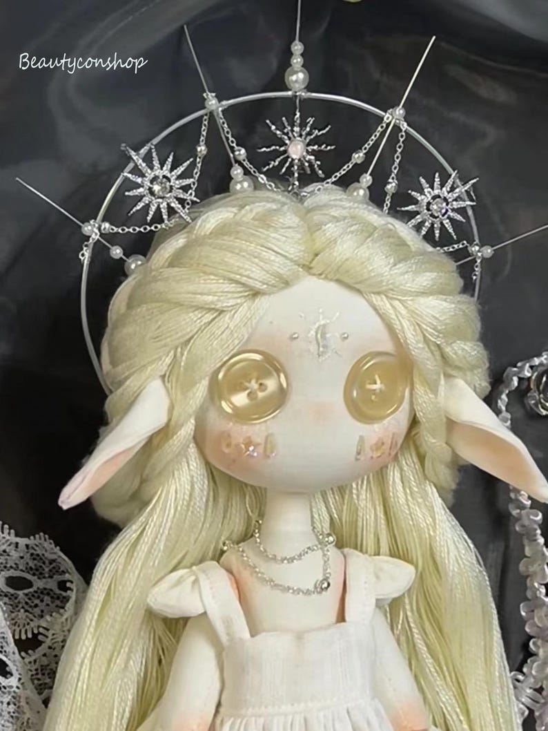 May include: A white porcelain doll with long blonde hair wearing a white dress and a silver crown with pearl accents. The doll has large, round, yellow button eyes.