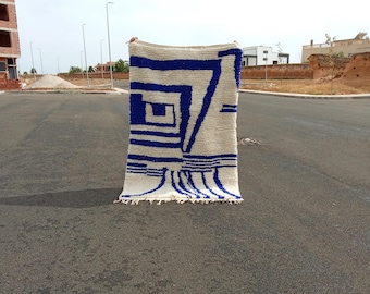 Puede incluir: Una alfombra rectangular de color blanquecino con un diseño geométrico en azul. La alfombra presenta una serie de líneas y formas azules, incluyendo cuadrados y rectángulos. La alfombra se sostiene frente a una carretera pavimentada.