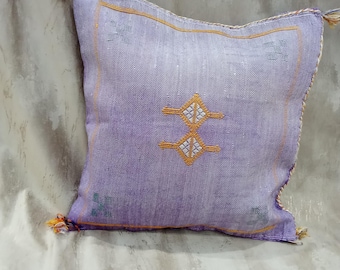 Puede incluir: Un cojín cuadrado lavanda con un diseño geométrico en hilo dorado y blanco. El cojín tiene ribetes dorados y borlas decorativas en cada esquina. El cojín está hecho de una tela texturizada.