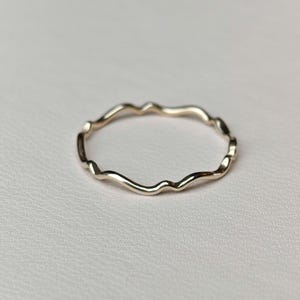 May include: A delicate gold ring with a wavy, organic design. The ring's surface reflects light, highlighting its flowing form. The ring is set against a neutral background, emphasising its elegant simplicity. A piece of jewellery.
