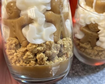May include: Two glass jars filled with a layered candle. The bottom layer is brown, the middle layer is a light brown crumble, the next layer is white frosting, and the top layer is gingerbread men.