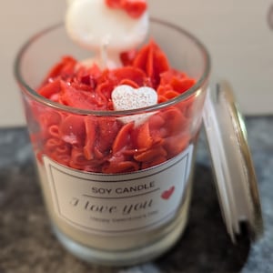 Puede incluir: Una vela de soja roja y blanca en un tarro de vidrio con una figurita blanca de Hello Kitty en la parte superior. La vela está decorada con corazones rojos y la etiqueta dice "Soy Candle I love you Happy Valentine's Day".