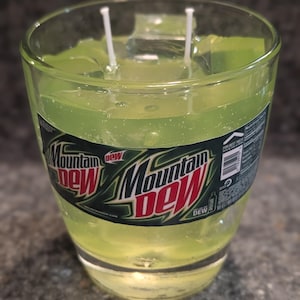 May include: A clear glass candle with a bright green wax interior. The candle features the Mountain Dew logo on the glass and two white wicks. The candle is a unique decorative item.