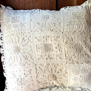 Może przedstawiać: Kremowa kwadratowa poduszka z patchworkowym wzorem. Poduszka posiada misterne koronki i haftowane detale, z falistym koronkowym wykończeniem na krawędziach. Projekt zawiera wzory kwiatowe i geometryczne.