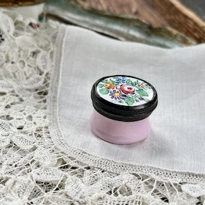 May include: A small, pink antique trinket box with a floral design on the lid. The lid is black with a white background and features a colourful arrangement of flowers. The box sits on a white lace doily.