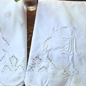 May include: Two white rectangular linens featuring intricate cutwork embroidery along the scalloped edges. One piece displays a prominent embroidered letter A centered within a circular floral wreath design, while the other features a smaller square motif.