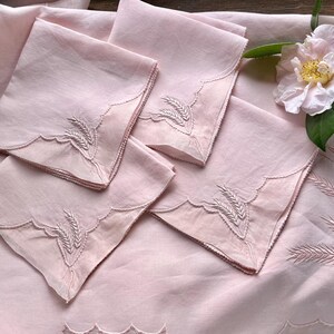 May include: A set of four square light pink napkins and a matching tablecloth, each featuring scalloped edges and embroidered wheat stalk motifs. A single pale pink camellia flower with green leaves rests on the fabric surface.