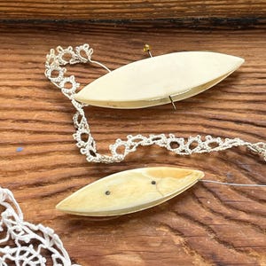 May include: Two off-white, boat-shaped tatting shuttles with small pins and thread, alongside a length of delicate, cream-colored lace. The shuttles rest on a warm, wooden surface, suggesting a craft or hobby theme.