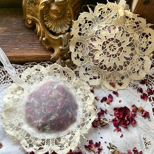 May include: Two handmade sachets filled with dried rose petals, presented on a white lace doily. The sachets are round, with a delicate lace trim. Scattered rose petals add a touch of color. A gold-colored ornate frame is in the background.