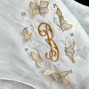 Puede incluir: Pañuelo blanco bordado con mariposas doradas y plateadas y detalles florales. Una letra monograma dorada "D" está centrada en el pañuelo. El delicado bordado añade un toque de elegancia a este accesorio vintage.